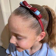 A young girl with her hair in a ponytail wearing a red headband with a light blue bow and a rhinestone center, paired with a light blue polo shirt.