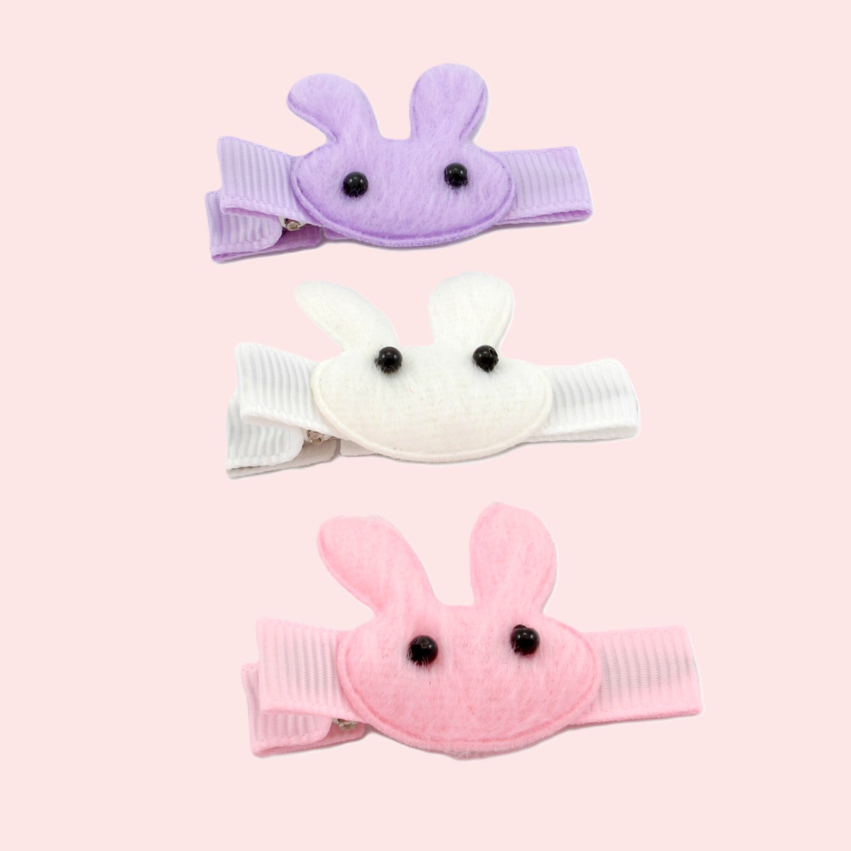 Three bunny-shaped hair clips in purple, white, and pink on a light pink background.