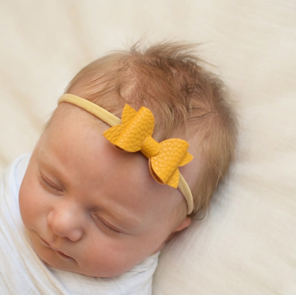 Newborn baby sleeping, wearing a mustard coloured leather bow headband 