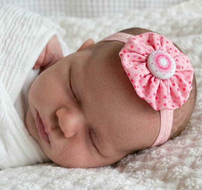 Newborn baby wearing a pink spotty yoyo flower headband and laying on a white blanket